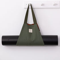 A green cotton MSY Mat Baguette with orange trim, hanging on a white hook with a black yoga mat rolled up inside it.