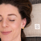A person with closed eyes rests their head on a beige Maya Shanti Yoga Pillow, made from sustainable hemp with ergonomic support and a small white label, placed on a textured dark surface.