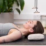 A woman lies on her back on a mat using the Maya Shanti Yoga Pillow for ergonomic head support. She is beneath hanging chimes, with plants and candles in the background.
