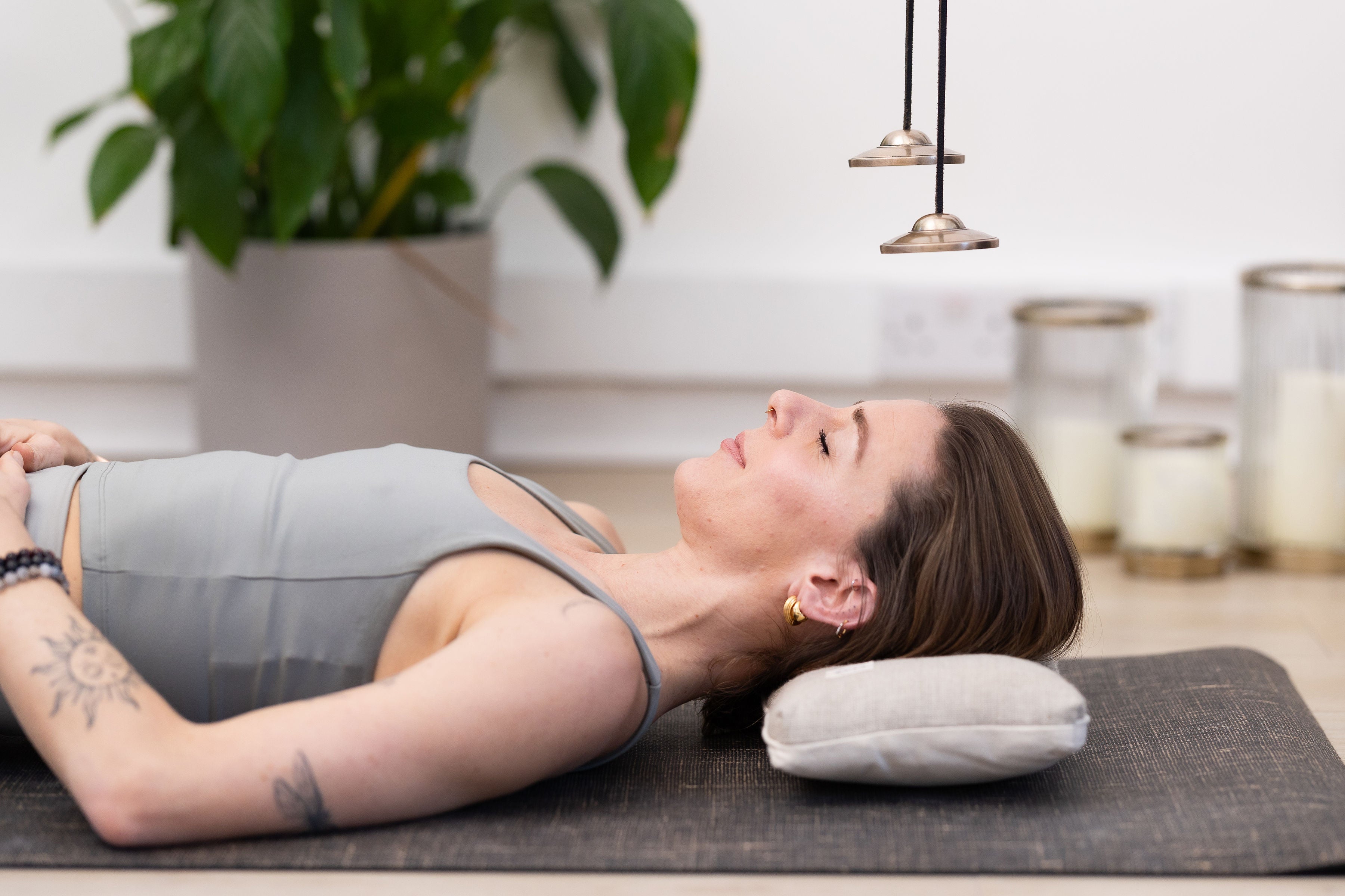 A woman lies on her back on a mat using the Maya Shanti Yoga Pillow for ergonomic head support. She is beneath hanging chimes, with plants and candles in the background.